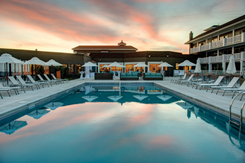 A serene pool area with lounge chairs and umbrellas, reflecting a colorful sunset and a nearby building.