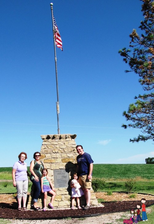 A family stands by a stone monument with an American flag against a clear blue sky. Green fields are in the background.