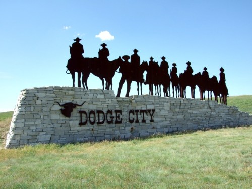 Silhouette of cowboys on horseback at the Dodge City welcome sign against a clear blue sky.
