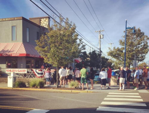 A crowd of people waiting outside Kohler's Bakery on a sunny day, with trees and power lines in the background.