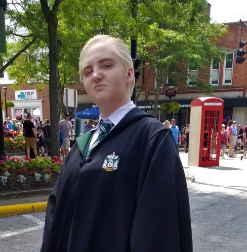 A person in a Slytherin robe stands confidently on a street, with shops and a red phone booth in the background.