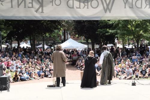 A crowd watches a performance on stage, with three actors in costume facing the audience under a sunny sky.