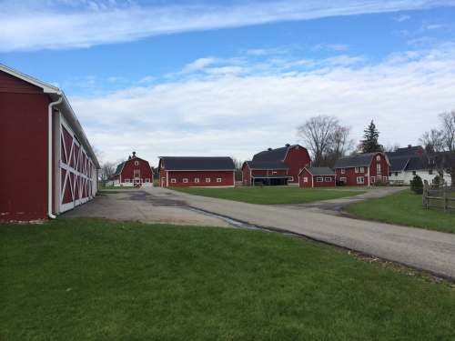 A scenic view of a red barn complex with a gravel road and green grass under a blue sky with clouds.