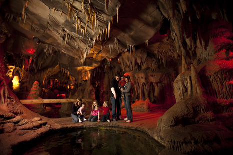 A family explores a cave with stalactites and stalagmites, gathered around a small pool of water. Soft red lighting enhances the scene.