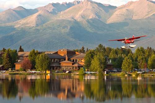 A seaplane flies over a lake, reflecting mountains and a lodge surrounded by trees in the background.