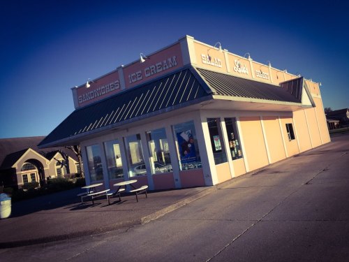 A pastel-colored building with signs for sandwiches and ice cream, featuring outdoor seating and large windows.