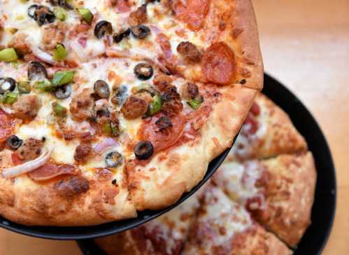 A close-up of a pepperoni and sausage pizza topped with olives and green peppers, with a second pizza in the background.