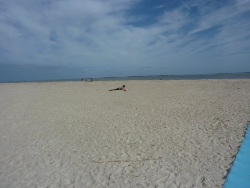 A wide sandy beach under a blue sky, with a few people in the distance and one person lying on the sand.