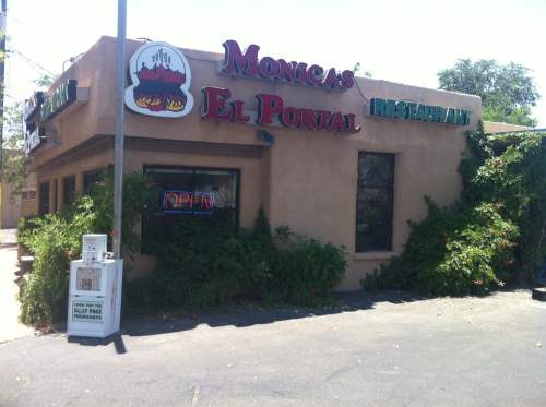 A restaurant building with a sign reading "Monica's El Portal," featuring an "Open" neon sign and surrounded by greenery.