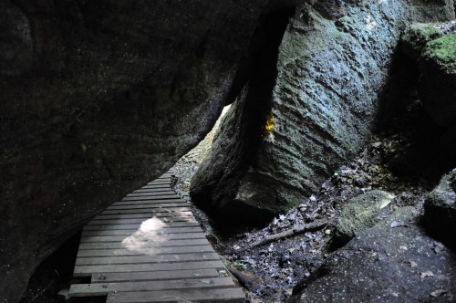 A narrow path made of wooden planks winds through large, moss-covered rocks in a shaded, natural setting.