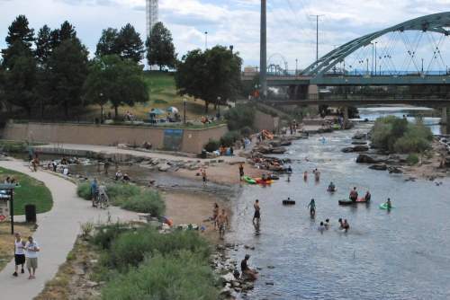 A river scene with people kayaking, swimming, and relaxing along the shore, surrounded by greenery and a bridge overhead.