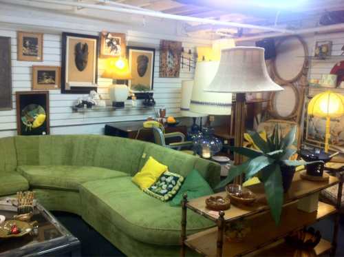 A cozy vintage shop interior featuring a green curved sofa, lamps, artwork, and decorative items on display.