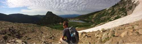 A hiker sits on rocky terrain, overlooking a scenic valley with a lake and mountains under a cloudy sky.