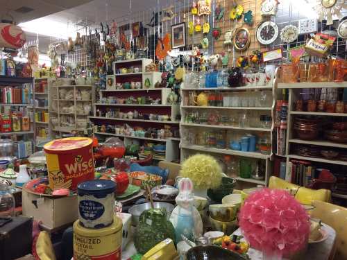 A cluttered vintage shop filled with colorful dishes, decor, and collectibles on shelves and tables.