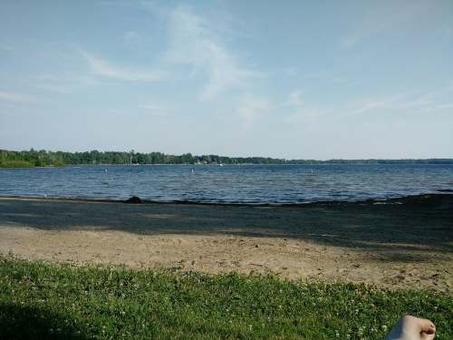 A serene lakeside view with sandy shore, green grass, and calm water under a clear blue sky.