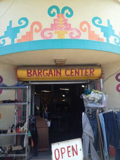 Entrance of a colorful store labeled "Bargain Center," with clothes and shoes displayed outside. Sign says "OPEN."