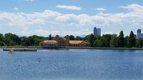A serene lake with a building in the background, surrounded by trees and a clear blue sky with fluffy clouds.