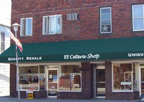 A brick storefront with a green awning reading "Et Cetera Shop," featuring large windows displaying various items for sale.