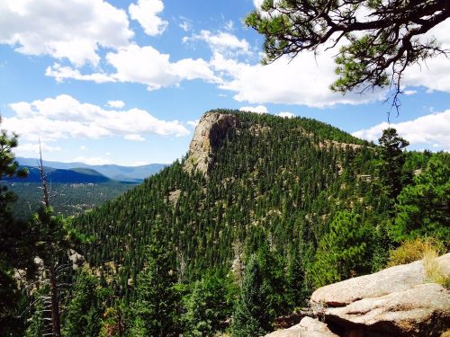 A scenic view of a forested mountain under a blue sky with fluffy clouds, showcasing rugged terrain and greenery.