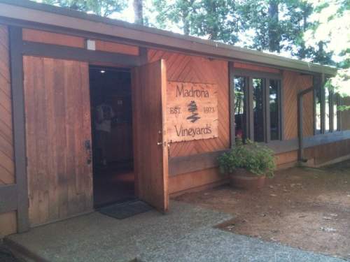 A wooden building with an open door, featuring a sign that reads "Makadonia Vineyards" surrounded by trees.