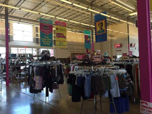 Interior of a thrift store with clothing racks and colorful banners hanging from the ceiling. Bright and spacious atmosphere.