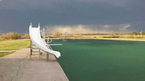 A swimming pool with a white slide, surrounded by green grass, under a cloudy sky.