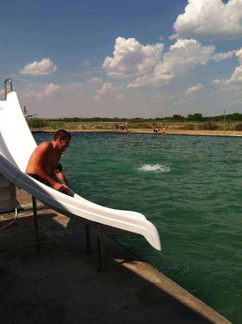 A shirtless man sits on a water slide, preparing to slide into a green pool under a partly cloudy sky.