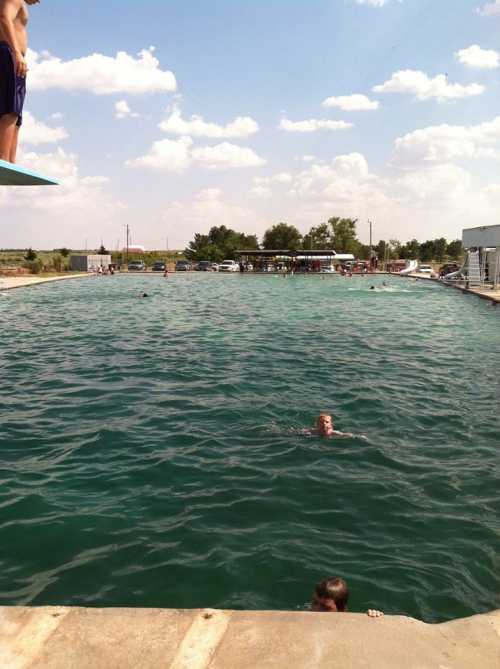 A swimming pool with people enjoying the water under a blue sky with clouds, surrounded by trees and buildings.