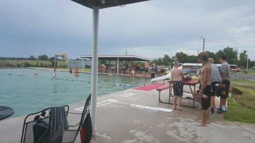 A pool scene with people swimming, playing basketball, and lounging by the water under a cloudy sky.