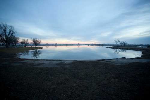 A calm lake at dusk, reflecting the cloudy sky and surrounding trees, with a grassy shore in the foreground.