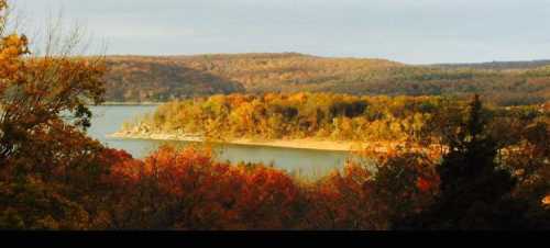 A scenic view of a lake surrounded by autumn foliage and rolling hills under a cloudy sky.