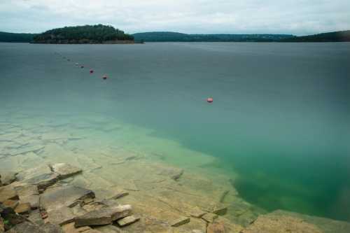 A calm lake with a rocky shore, featuring a gradient from clear water to a cloudy sky in the background.