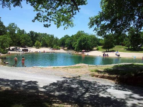 A serene pond surrounded by trees, with people enjoying the beach area and swimming on a sunny day.