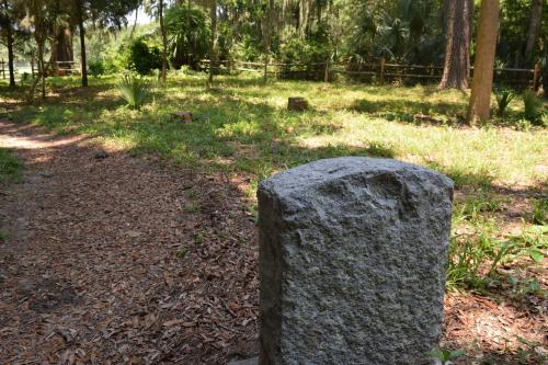 A weathered stone marker stands on a grassy path surrounded by trees and foliage in a serene outdoor setting.