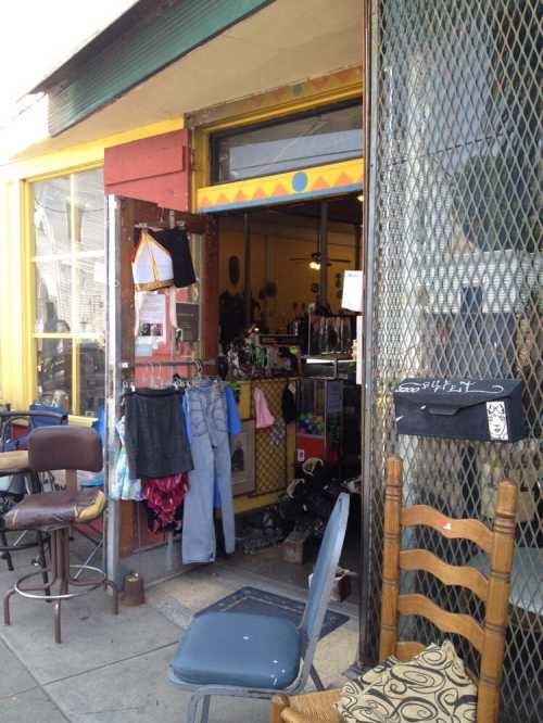 A colorful storefront with an open door, displaying clothing and accessories, surrounded by outdoor seating.