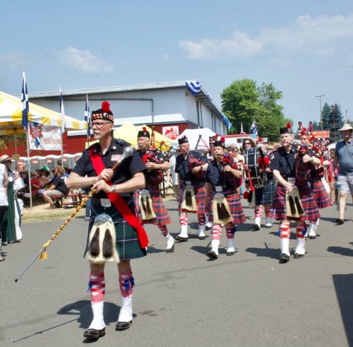 A marching band in kilts plays bagpipes during a parade, with colorful tents and spectators in the background.