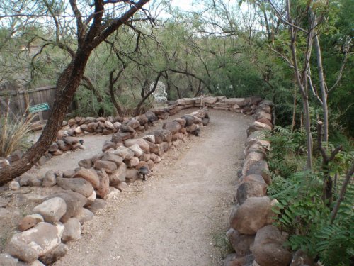 A winding dirt path bordered by stone walls, surrounded by greenery and trees in a natural setting.