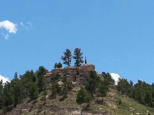 A rocky hill topped with trees under a clear blue sky, featuring a tall, slender structure on the summit.