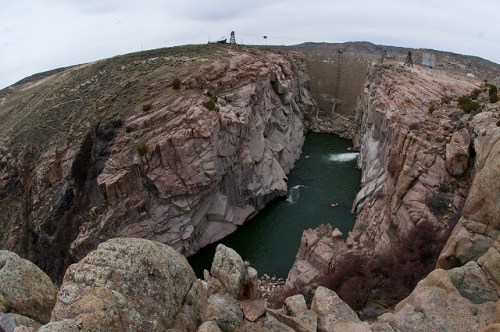 A panoramic view of a rocky canyon with a river at the bottom and a dam in the background under a cloudy sky.