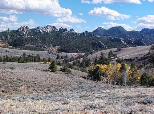 A scenic landscape featuring rolling hills, rocky formations, and scattered trees under a blue sky with fluffy clouds.