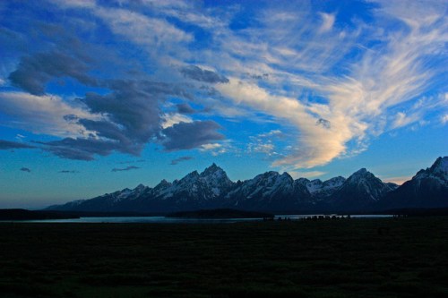 A scenic view of snow-capped mountains under a blue sky with wispy clouds, reflecting on a calm lake.