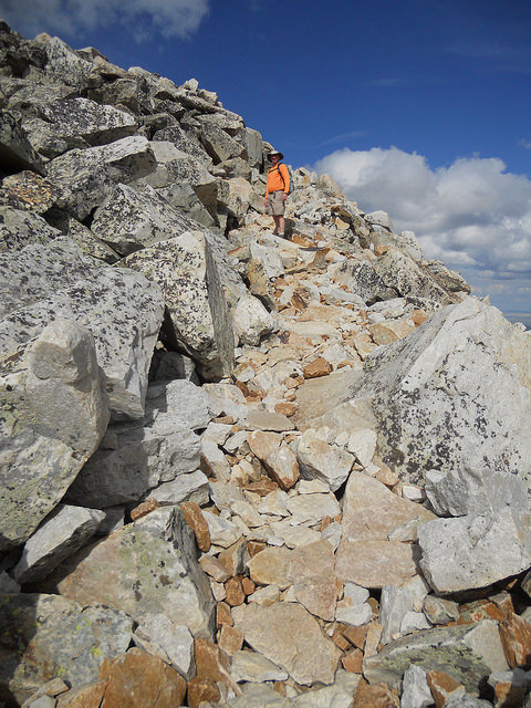 A hiker in an orange shirt navigates a rocky trail on a mountain under a blue sky with clouds.