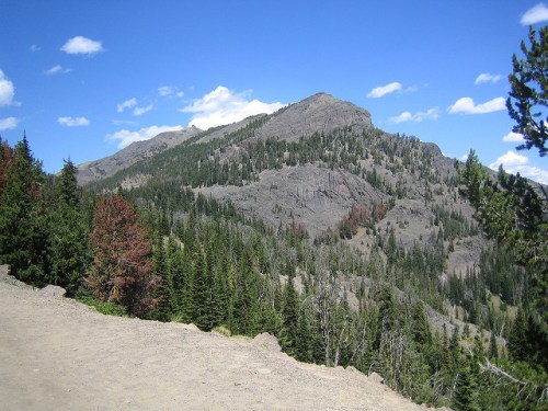 A scenic view of a mountain surrounded by lush green trees under a clear blue sky with scattered clouds.
