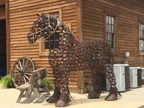 A large sculpture of a horse made from intertwined metal pieces, set against a wooden building backdrop.