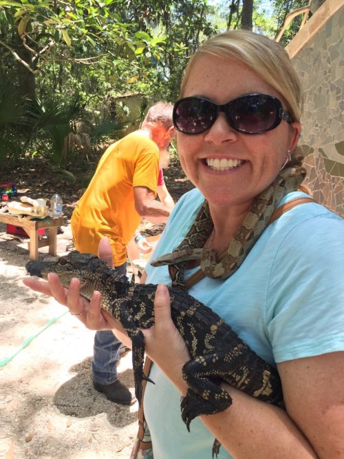 A woman smiles while holding a small alligator, with a snake draped around her neck in a lush outdoor setting.