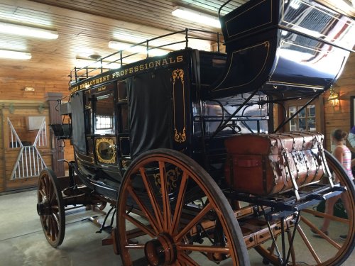 A vintage horse-drawn carriage with wooden wheels and a black exterior, displayed indoors with wooden walls.