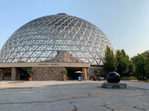 A large glass dome structure with a stone entrance, surrounded by trees and a black spherical sculpture in the foreground.