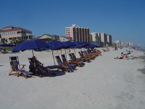A beach scene with blue umbrellas and lounge chairs, people relaxing on the sand, and buildings in the background.