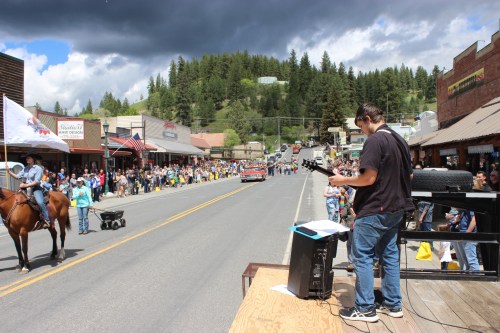 A parade scene with a crowd lining the street, a boy on a platform, and a horse passing by in a small town.