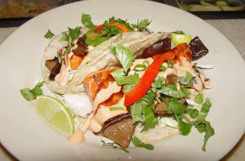 Two tacos filled with grilled meat, topped with red peppers, cilantro, and a creamy sauce, served with lime wedges.